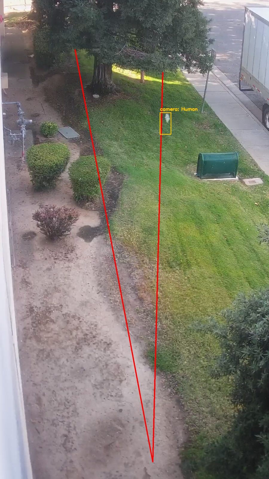 A grass verge beside a path, with a
    small pale object on the lawn marked by an amber box labelled camera Human,
    next to a green utility cabinet for scale