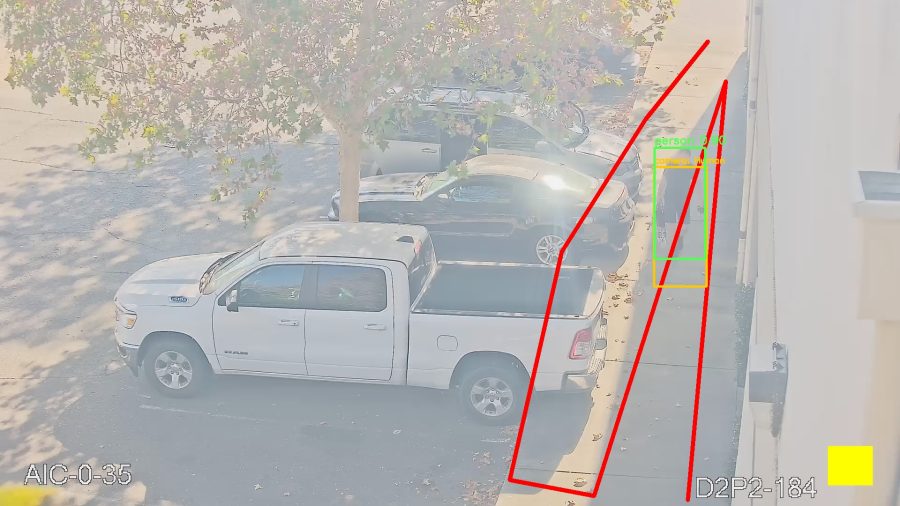 A pedestrian walking along a pavement
    beside parked vehicles, boxed in green as 'person 0.90' with the camera's
    amber box slightly lower